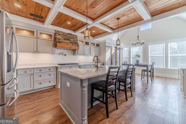 kitchen with appliances with stainless steel finishes, hanging light fixtures, wood ceiling, light stone counters, and a center island with sink