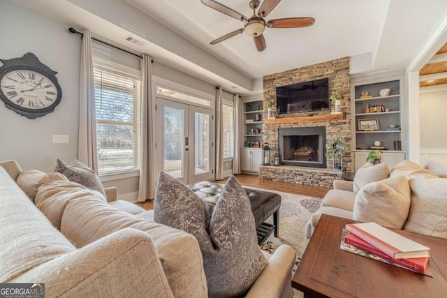 living room featuring a stone fireplace, built in features, hardwood / wood-style flooring, ceiling fan, and french doors