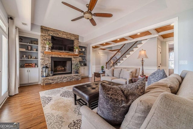 living room featuring built in features, coffered ceiling, a stone fireplace, beamed ceiling, and light wood-type flooring