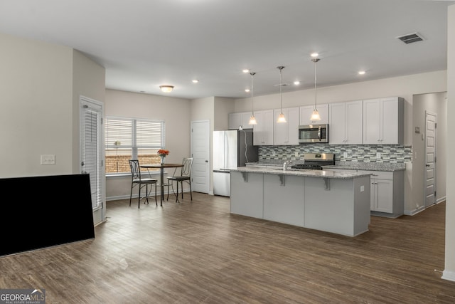 kitchen with pendant lighting, dark wood-type flooring, appliances with stainless steel finishes, light stone counters, and a center island with sink