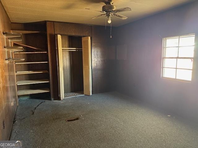 unfurnished bedroom with ceiling fan, wooden walls, and dark colored carpet