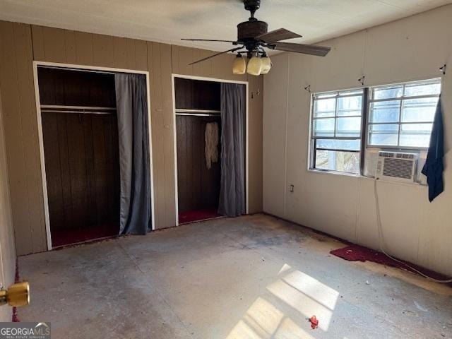 unfurnished bedroom with cooling unit, two closets, wooden walls, and ceiling fan