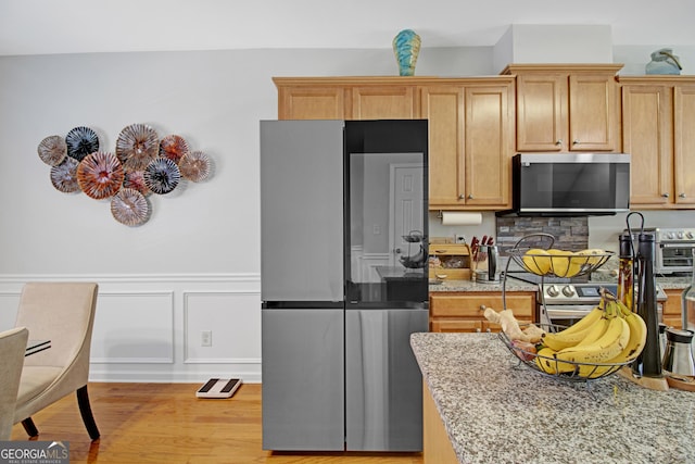 kitchen with light stone counters, a wainscoted wall, light wood finished floors, a decorative wall, and appliances with stainless steel finishes