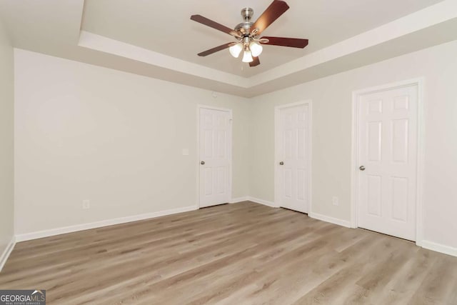 unfurnished bedroom featuring two closets, light hardwood / wood-style flooring, ceiling fan, and a tray ceiling