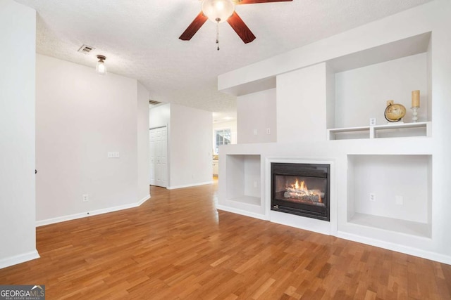 unfurnished living room with ceiling fan, built in shelves, wood-type flooring, and a textured ceiling