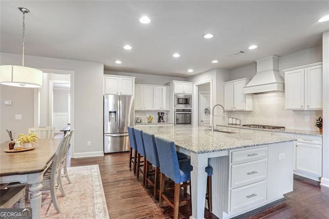 kitchen with sink, premium range hood, appliances with stainless steel finishes, an island with sink, and white cabinets