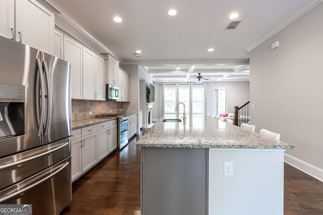 kitchen with coffered ceiling, sink, light stone counters, appliances with stainless steel finishes, and an island with sink