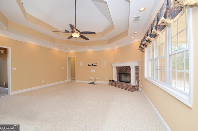 unfurnished living room featuring carpet flooring, ceiling fan, a tray ceiling, crown molding, and a brick fireplace