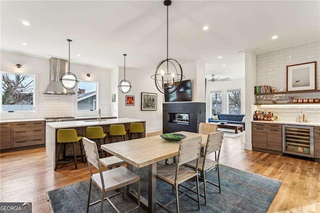 dining area with a multi sided fireplace, ornamental molding, wet bar, beverage cooler, and light wood-type flooring