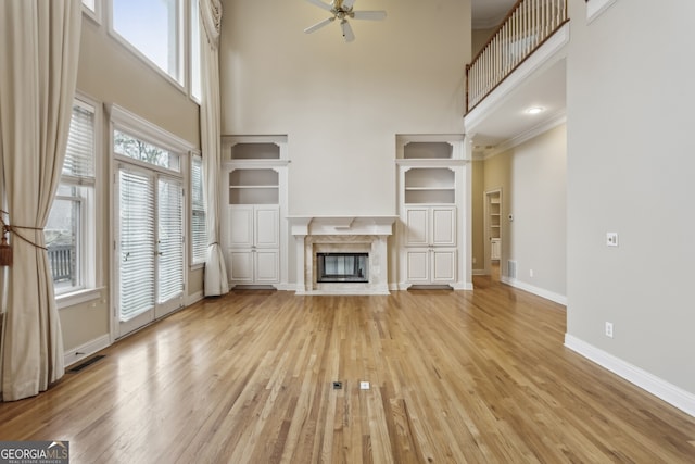 unfurnished living room featuring built in features, ceiling fan, a premium fireplace, light hardwood / wood-style floors, and a high ceiling