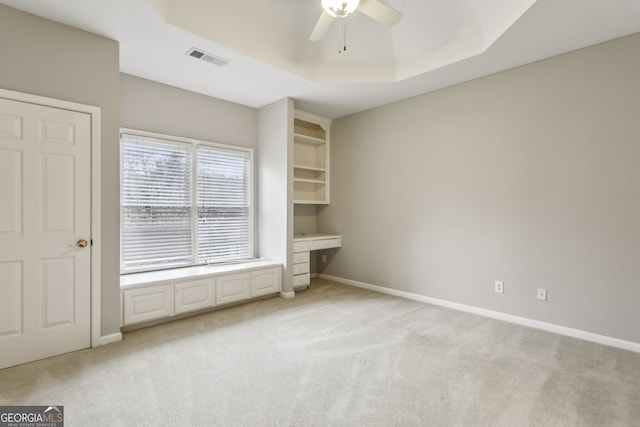 unfurnished bedroom with a raised ceiling, built in desk, light colored carpet, and ceiling fan