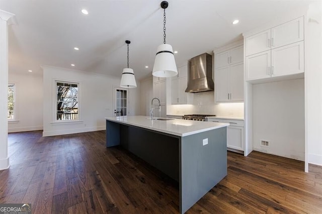 kitchen featuring sink, decorative light fixtures, a center island with sink, wall chimney range hood, and white cabinets