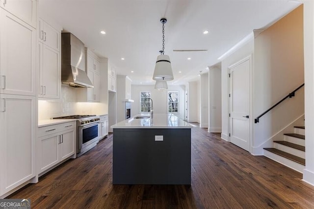 kitchen with stainless steel stove, white cabinetry, hanging light fixtures, wall chimney range hood, and a center island with sink