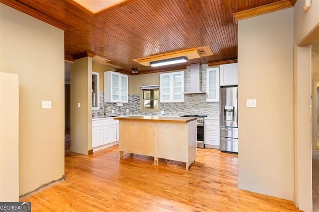 kitchen with wall chimney range hood, appliances with stainless steel finishes, white cabinetry, tasteful backsplash, and a kitchen island