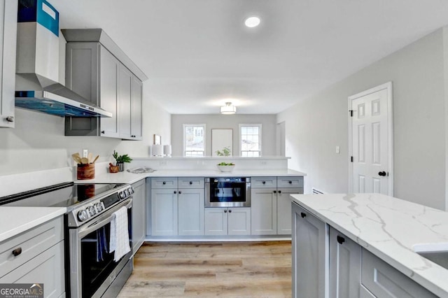 kitchen featuring light hardwood / wood-style flooring, gray cabinets, appliances with stainless steel finishes, light stone countertops, and wall chimney exhaust hood