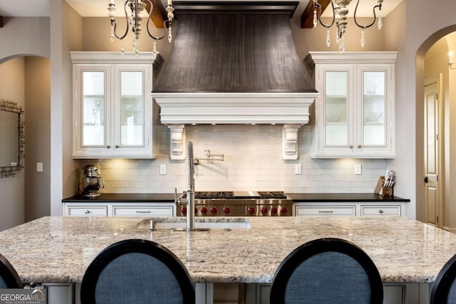 kitchen with an island with sink, premium range hood, glass insert cabinets, and dark stone counters