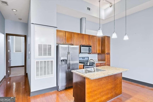 kitchen with an island with sink, hanging light fixtures, light stone counters, light hardwood / wood-style floors, and stainless steel appliances