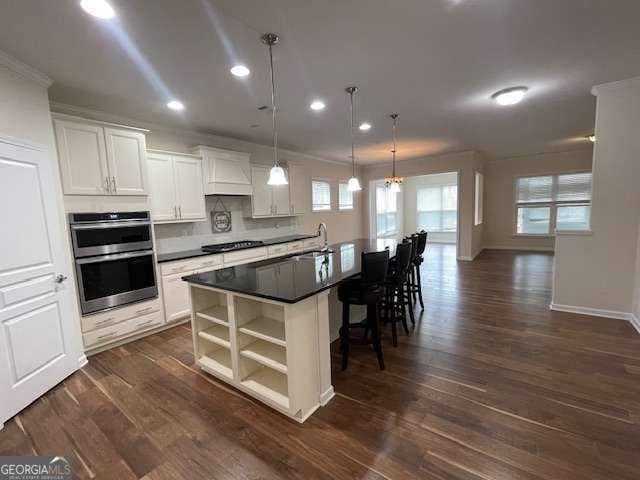 kitchen with double oven, decorative light fixtures, white cabinetry, an island with sink, and wall chimney exhaust hood