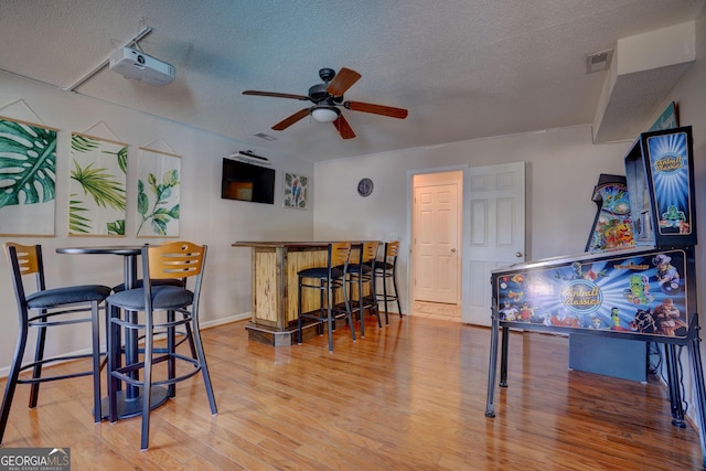 rec room with ceiling fan, a textured ceiling, bar area, and light hardwood / wood-style floors