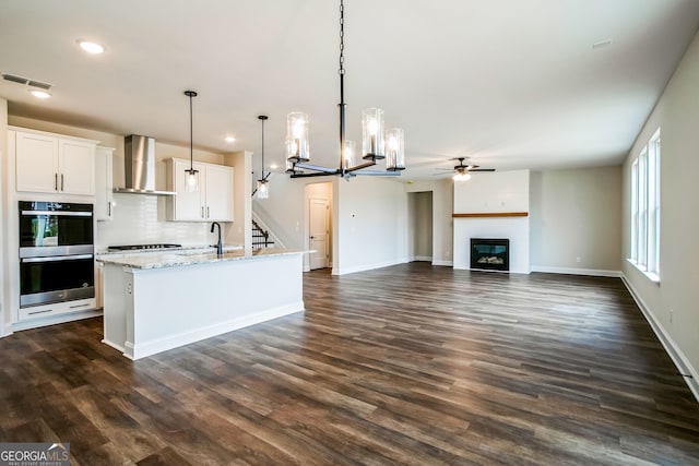 kitchen with wall chimney range hood, hanging light fixtures, an island with sink, white cabinets, and stainless steel double oven