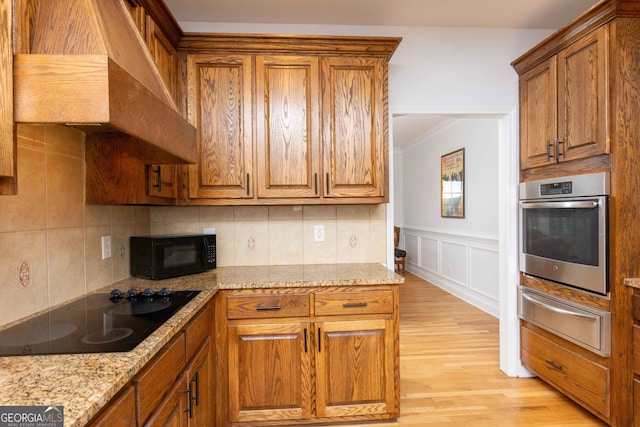 kitchen featuring wall chimney range hood, backsplash, light stone counters, black appliances, and light wood-type flooring