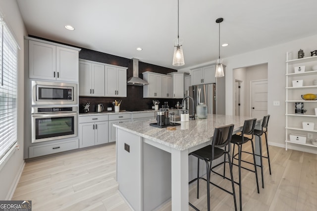 kitchen with wall chimney exhaust hood, decorative light fixtures, appliances with stainless steel finishes, a kitchen breakfast bar, and an island with sink