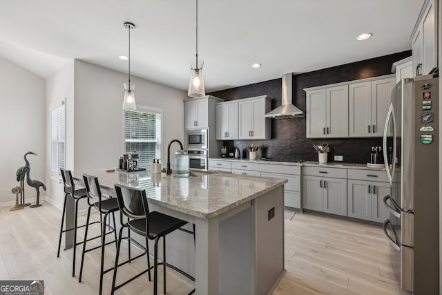 kitchen with wall chimney range hood, appliances with stainless steel finishes, hanging light fixtures, light stone countertops, and an island with sink