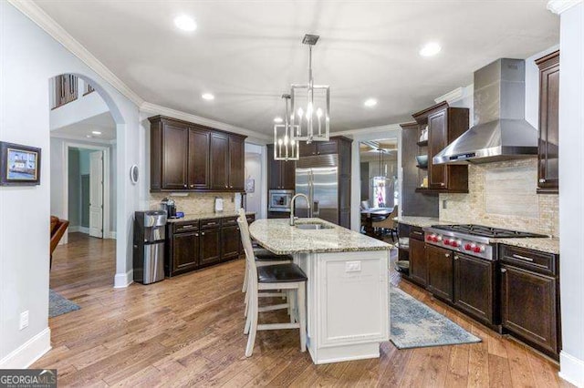 kitchen with an island with sink, sink, built in appliances, light stone counters, and wall chimney range hood