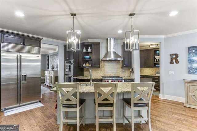 kitchen with an island with sink, dark brown cabinetry, stainless steel appliances, light stone countertops, and wall chimney exhaust hood