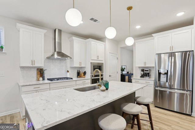 kitchen with hanging light fixtures, wall chimney range hood, an island with sink, stainless steel appliances, and white cabinets