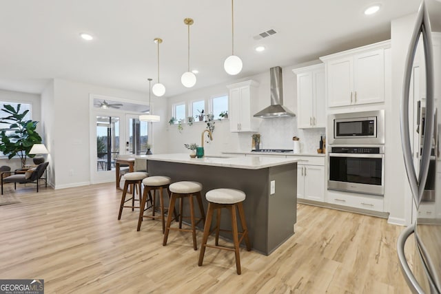 kitchen featuring white cabinetry, stainless steel appliances, a center island with sink, decorative light fixtures, and wall chimney exhaust hood