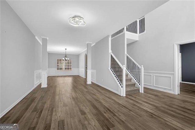 unfurnished living room featuring dark wood-style flooring, crown molding, a decorative wall, stairway, and a chandelier