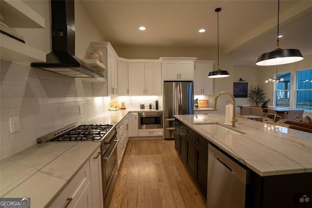 kitchen with wall chimney range hood, sink, white cabinetry, a kitchen island with sink, and stainless steel appliances