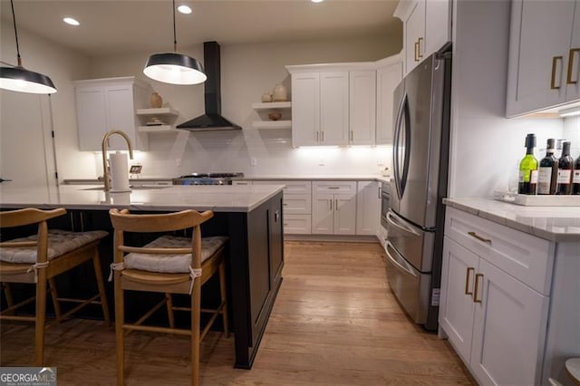 kitchen with wall chimney exhaust hood, stainless steel fridge, a kitchen island with sink, and white cabinets