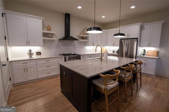 kitchen with wall chimney exhaust hood, a kitchen bar, a center island with sink, stainless steel appliances, and white cabinets