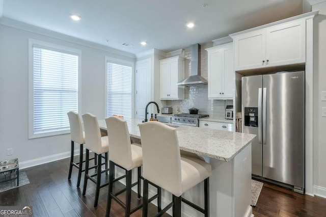 kitchen featuring a center island with sink, stainless steel refrigerator with ice dispenser, wall chimney range hood, and white cabinetry