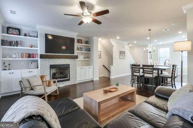 living room featuring a stone fireplace, ceiling fan with notable chandelier, ornamental molding, dark wood-type flooring, and built in shelves