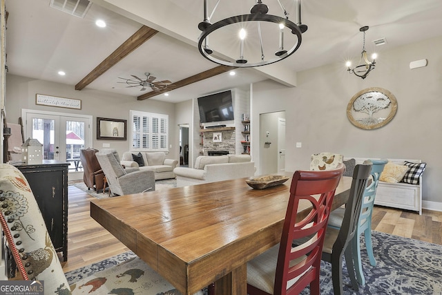 dining room featuring french doors, a stone fireplace, light wood-type flooring, beam ceiling, and ceiling fan with notable chandelier