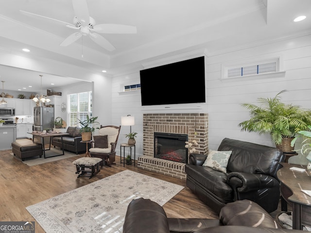 living room with a fireplace, ceiling fan with notable chandelier, wood-type flooring, and a raised ceiling