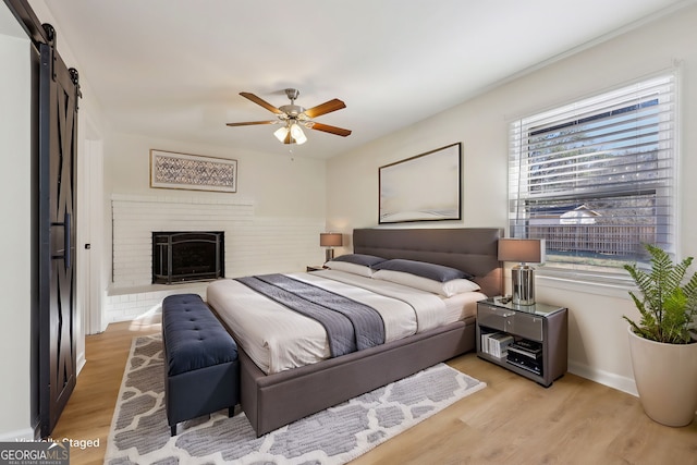 bedroom featuring a brick fireplace, light hardwood / wood-style flooring, a barn door, and ceiling fan