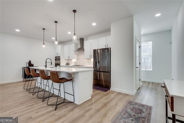 kitchen with appliances with stainless steel finishes, white cabinetry, a center island with sink, decorative light fixtures, and wall chimney exhaust hood