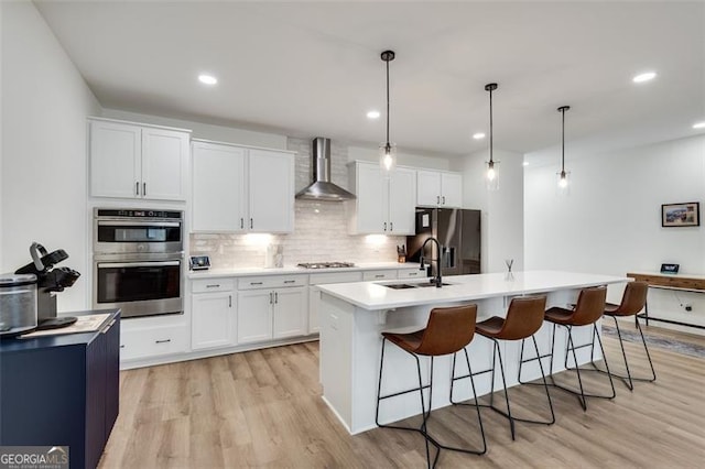 kitchen with wall chimney exhaust hood, hanging light fixtures, stainless steel appliances, a kitchen island with sink, and white cabinets