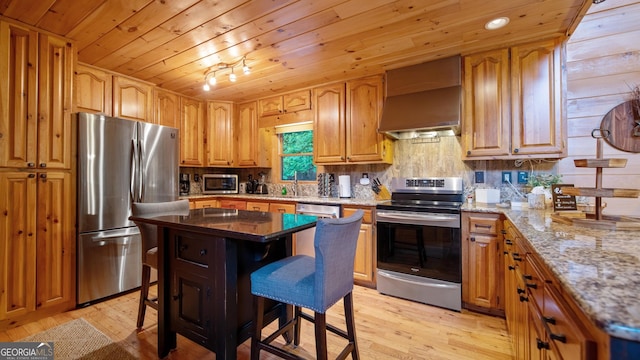 kitchen with a kitchen island, appliances with stainless steel finishes, a kitchen bar, light stone countertops, and wall chimney range hood