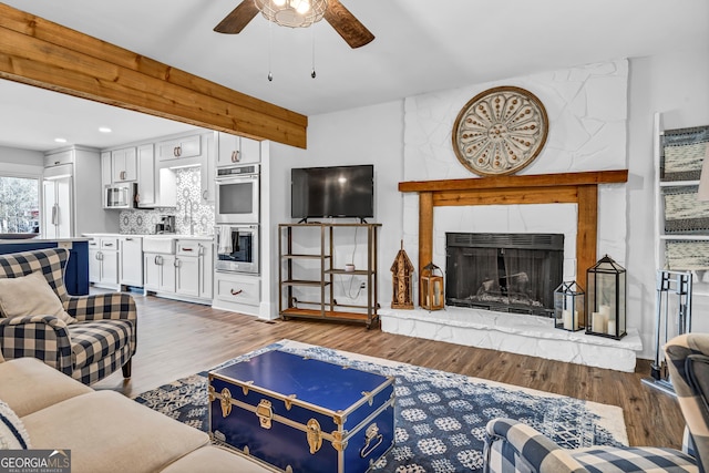 living room with ceiling fan, a large fireplace, beam ceiling, and light hardwood / wood-style flooring