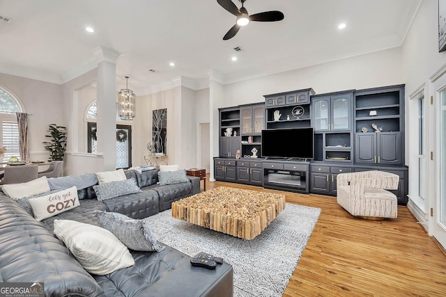 living room with decorative columns, visible vents, light wood-style floors, ornamental molding, and ceiling fan with notable chandelier