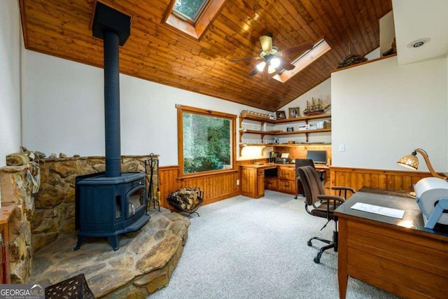 home office with vaulted ceiling with skylight, a wood stove, ceiling fan, light carpet, and wooden ceiling