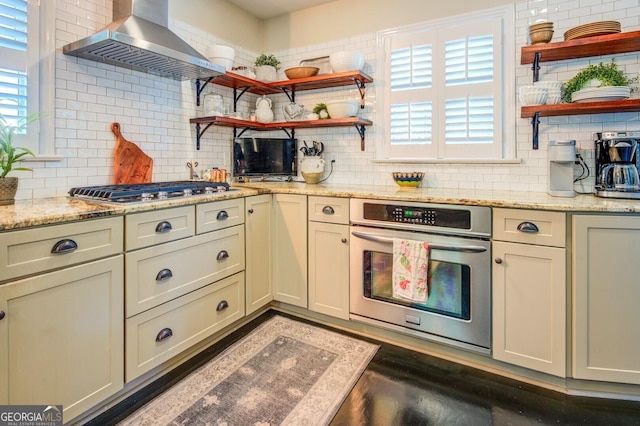 kitchen featuring light stone counters, appliances with stainless steel finishes, range hood, cream cabinets, and decorative backsplash