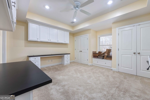 unfurnished office featuring light carpet, a tray ceiling, built in desk, and ceiling fan