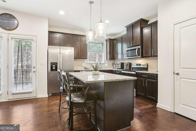 kitchen featuring dark brown cabinetry, hanging light fixtures, a kitchen island, stainless steel appliances, and light stone countertops