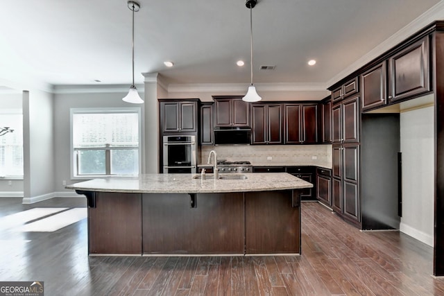 kitchen with sink, hanging light fixtures, dark brown cabinetry, light stone countertops, and an island with sink